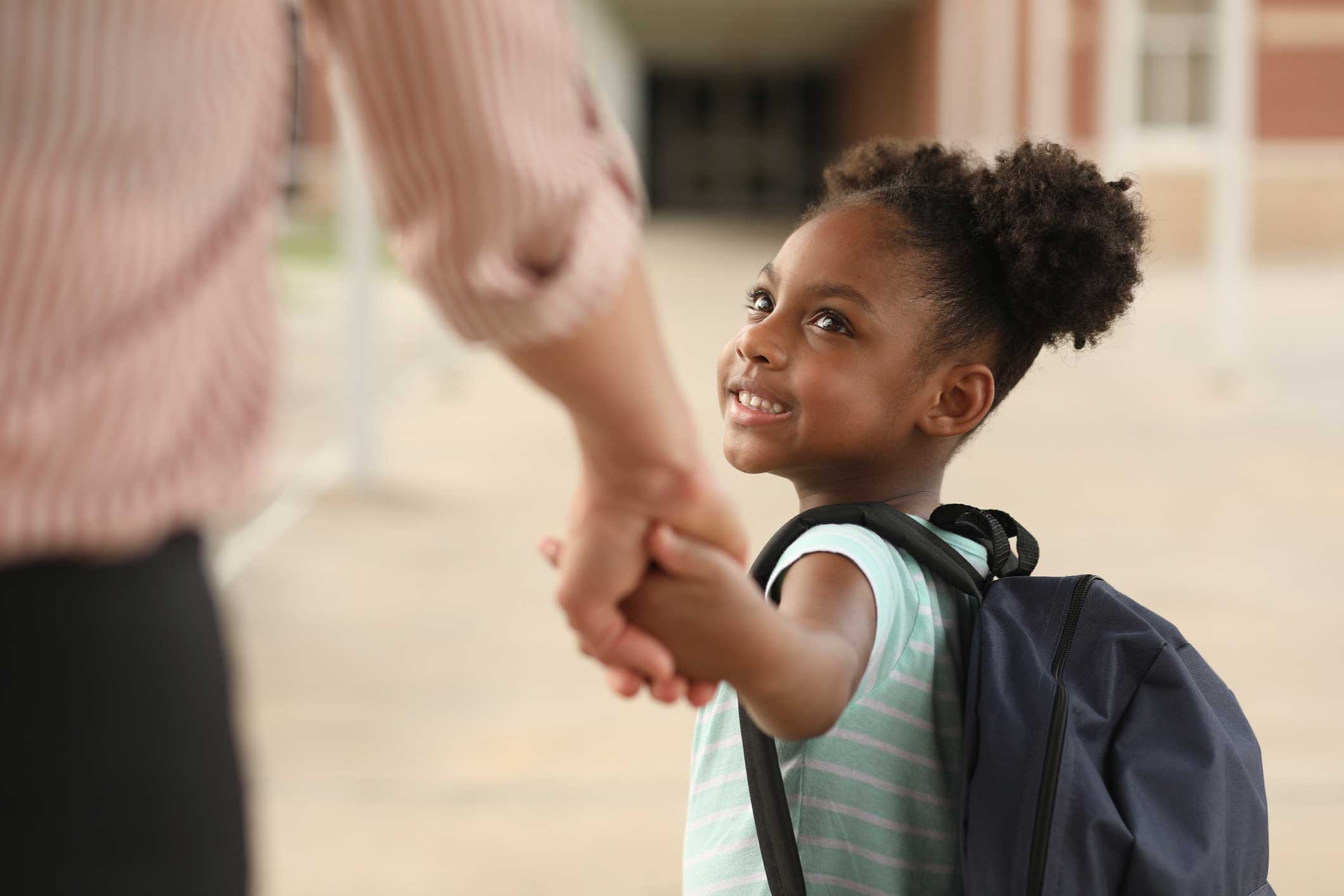 Black child in a backpack holding white parent's hand and smiling