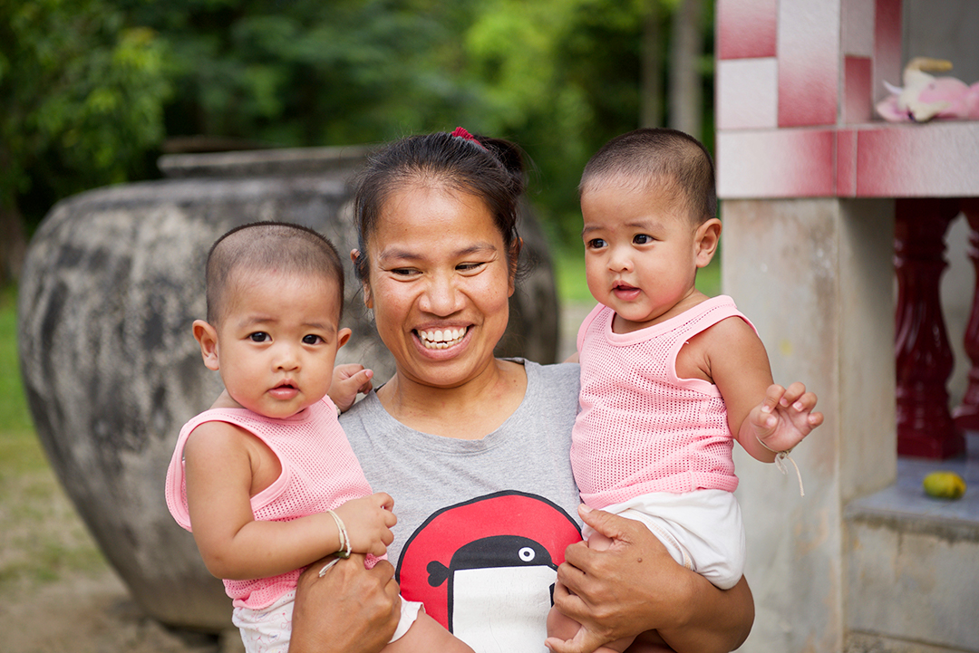 Ping holding her twins and smiling. When you sponsor a child in Thailand you help people like Ping.