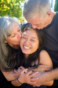 A photo of Adult adoptee Ying Lamb with her adopted parents, them kissing her on the head. She shares her advice for helping adopted children.