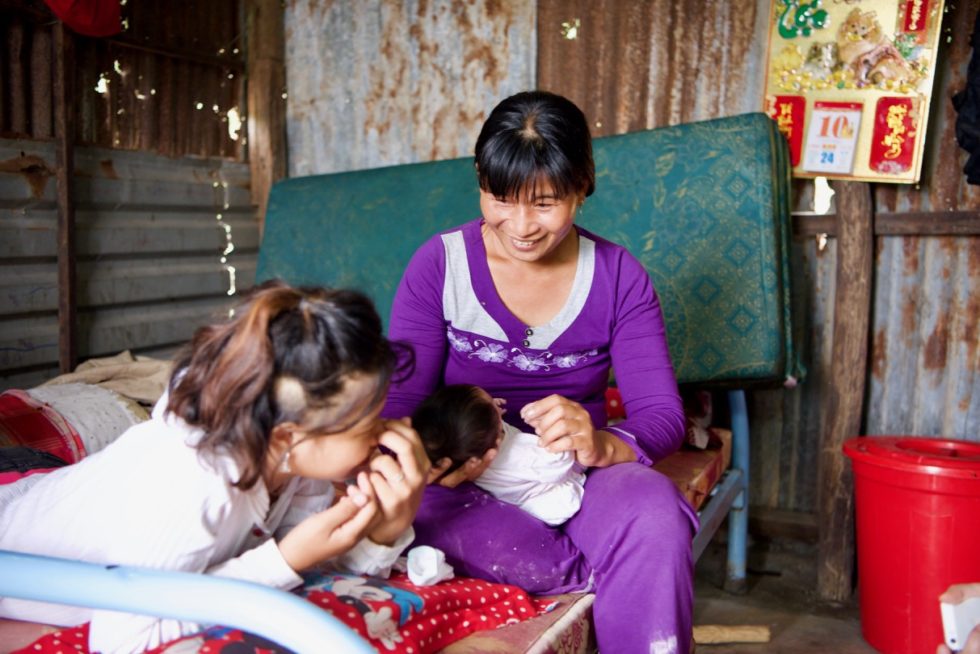 Linh lays on the bed while her mom smiles and holds Linh's baby. 