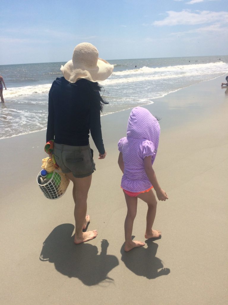Adoptive mom wearing a hat and shorts walking on the beach near the water alongside her adoptive child wearing a swimsuit and a purple hoodie