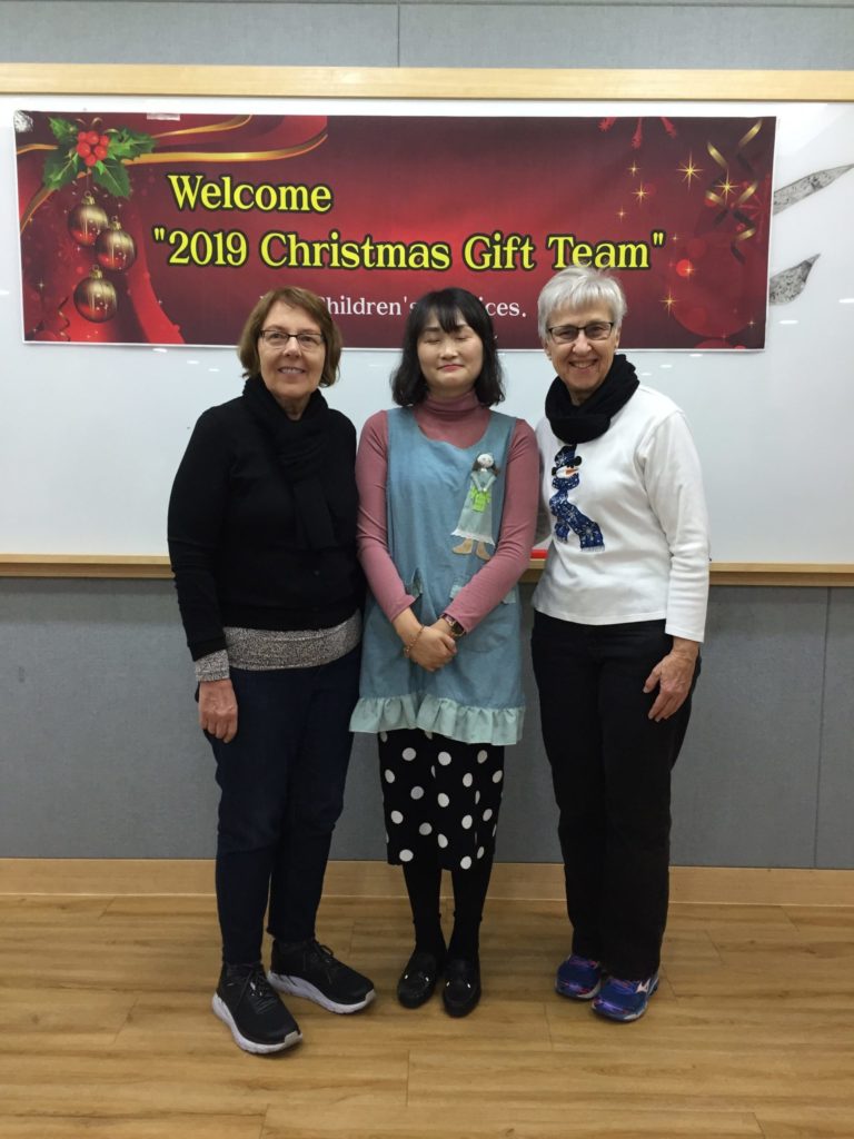 Seri (far right) and a fellow gift team member stand on either side of a calligraphy and art instructor at Holt Morning Garden shelter. 