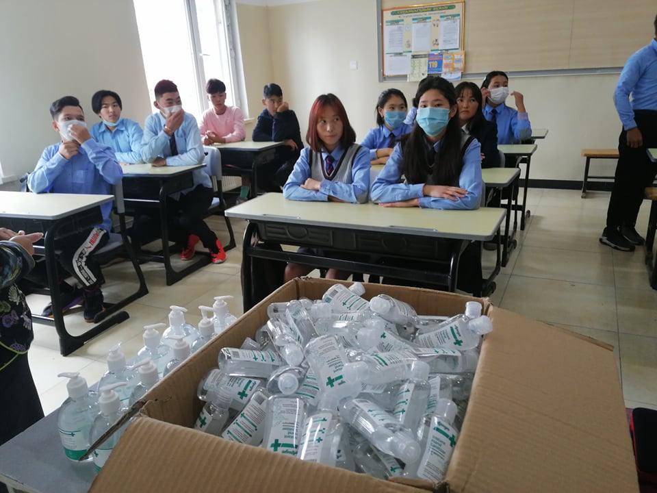 students in masks sitting at desks behind box of hand sanitizer