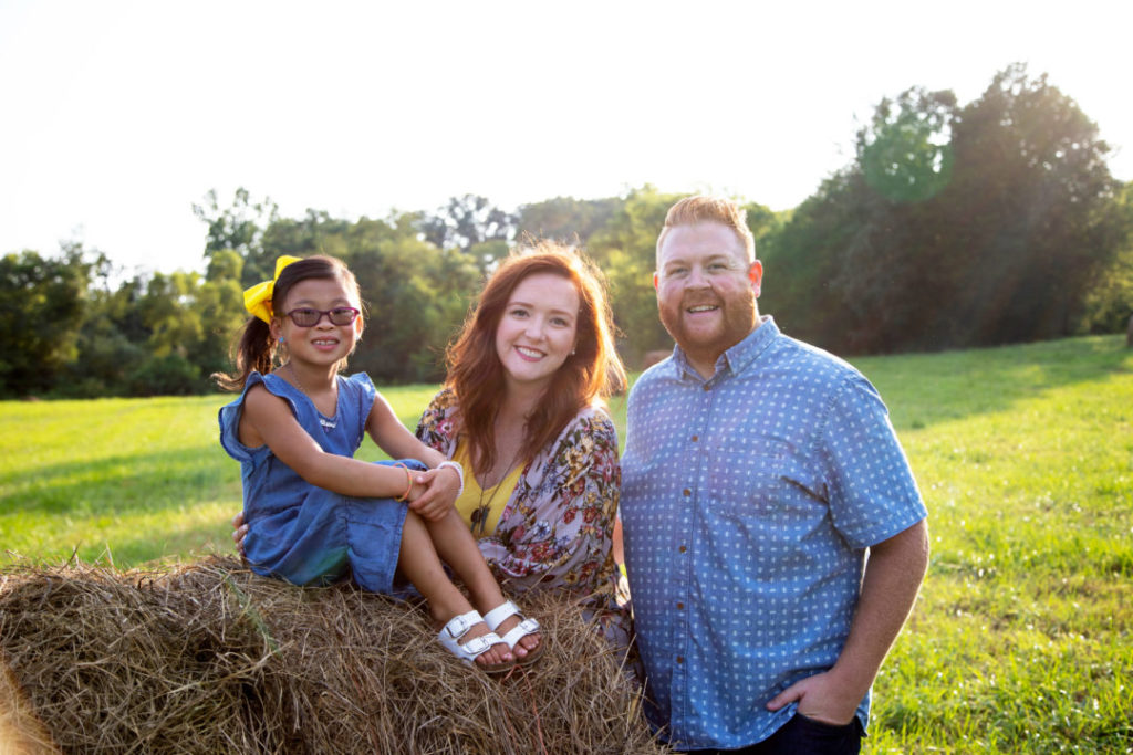 little girl sitting on haybale next to smiling mom and dad
