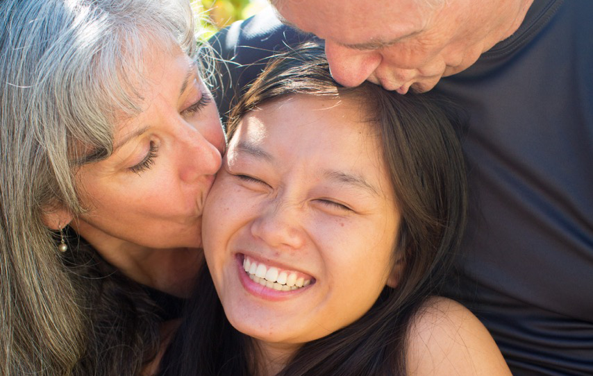 Adult adoptee Ying Lamb with her adopted parents, them kissing her on the head. She shares her advice for helping adopted children.
