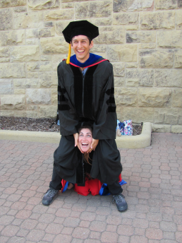 laughing man wearing graduation gown pretending to sit on laughing woman