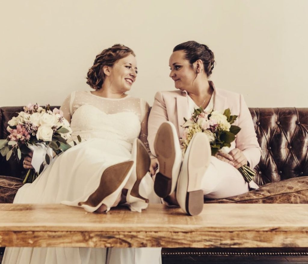 one woman in a wedding dress smiling at one woman in a wedding suit