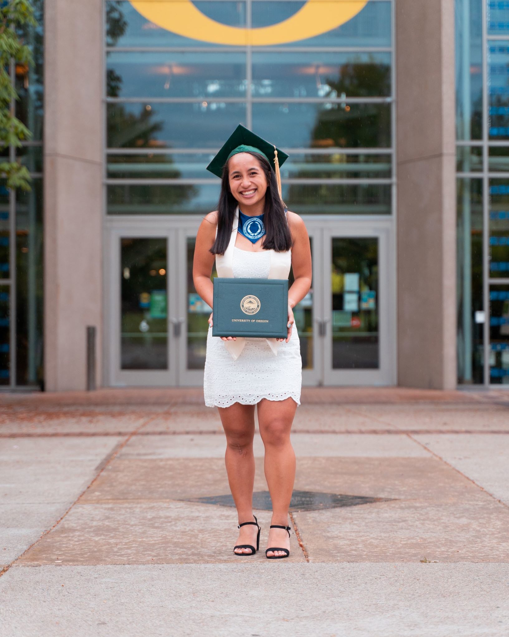 smiling girl wearing green graduation cap and white dress holding diploma in front of big yellow O