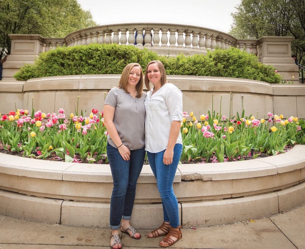two woman smiling standing in front of a bed of tulips