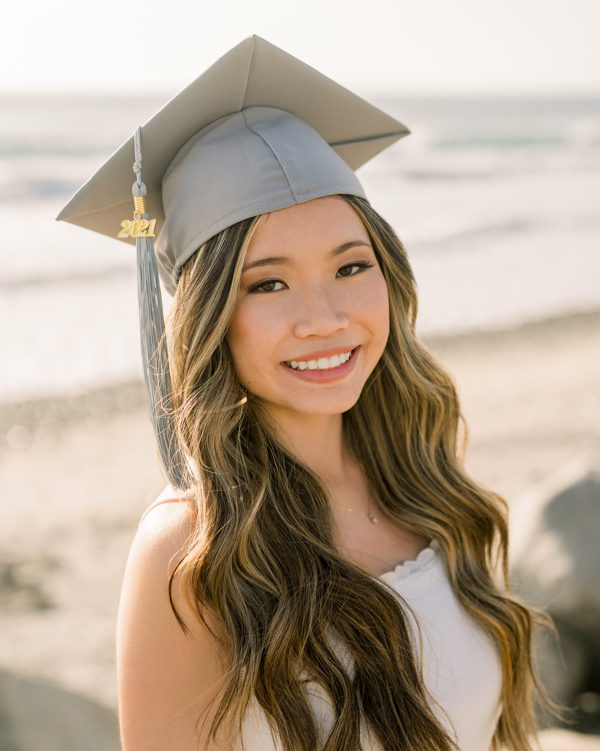 smiling girl on the beach with long wavy hair wearing grey graduation cap and white dress