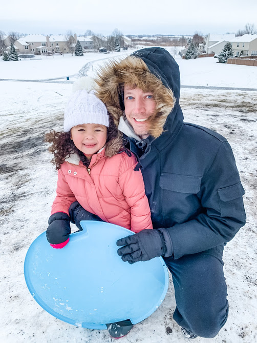 man holding little girl in pink fluffy coat and blue sled