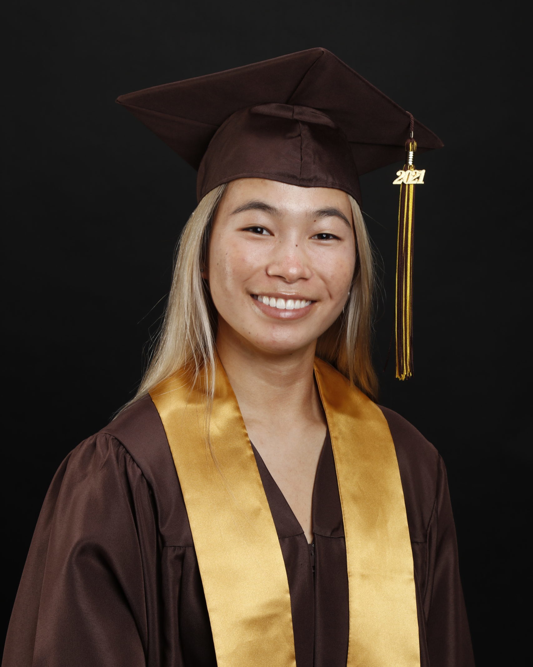 smiling girl in brown graduation cap and gown and yellow sash