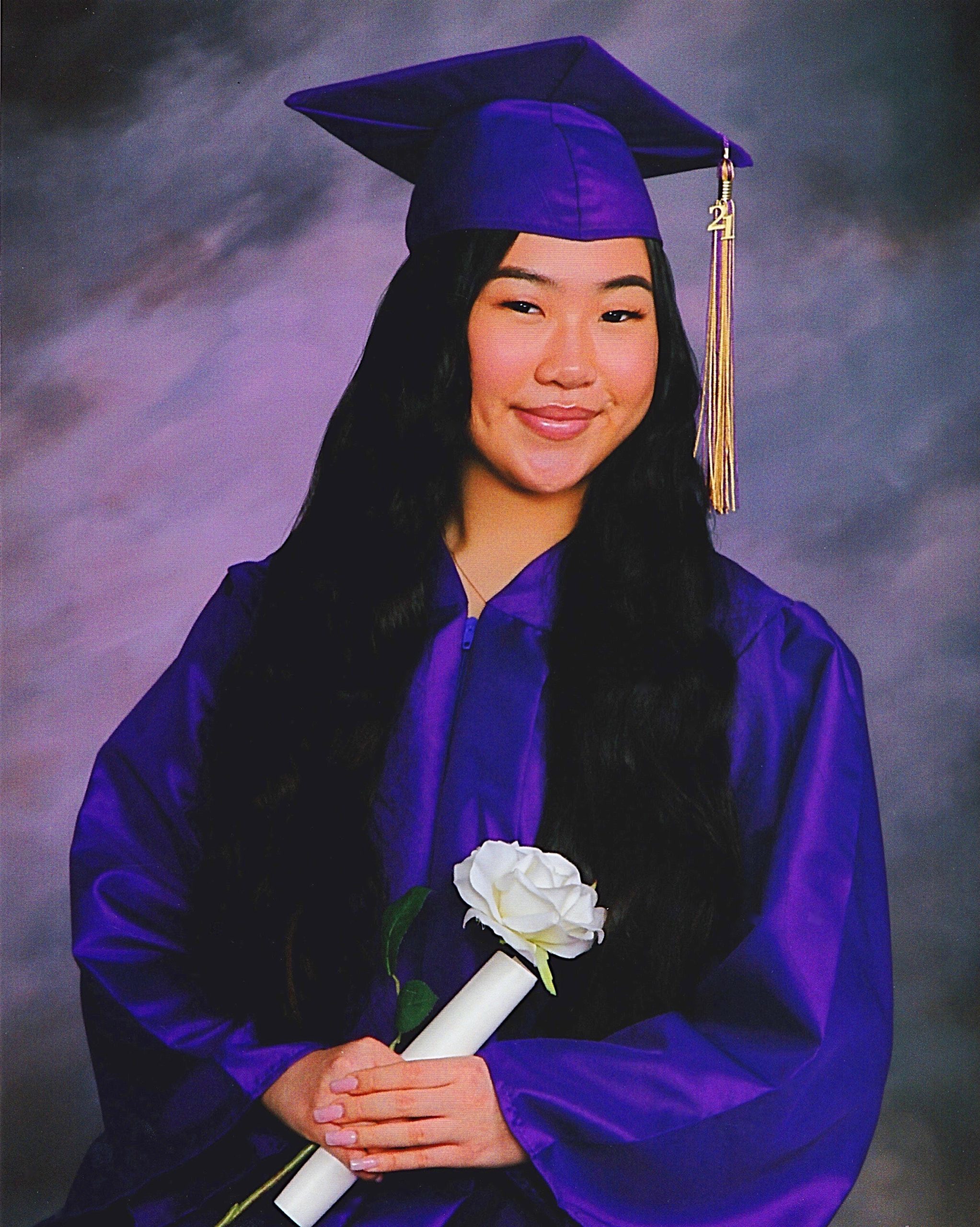 smiling girl wearing purple graduation cap and gown and holding white rose and rolled up diploma