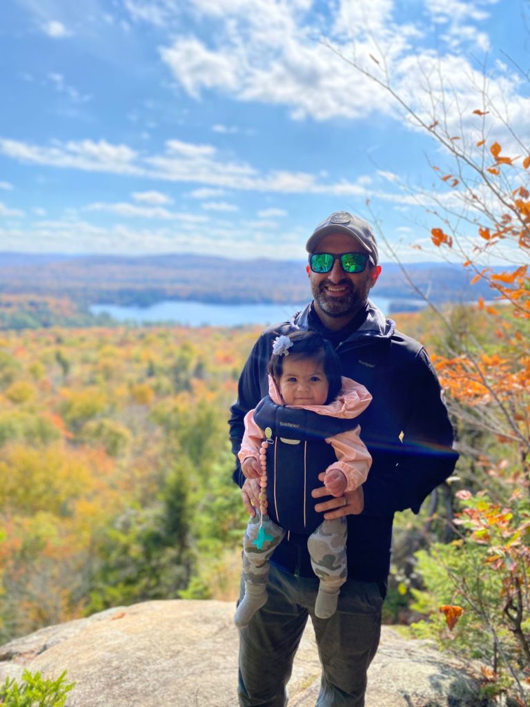 smiling man at top of hill wearing baby carrier with toddler girl
