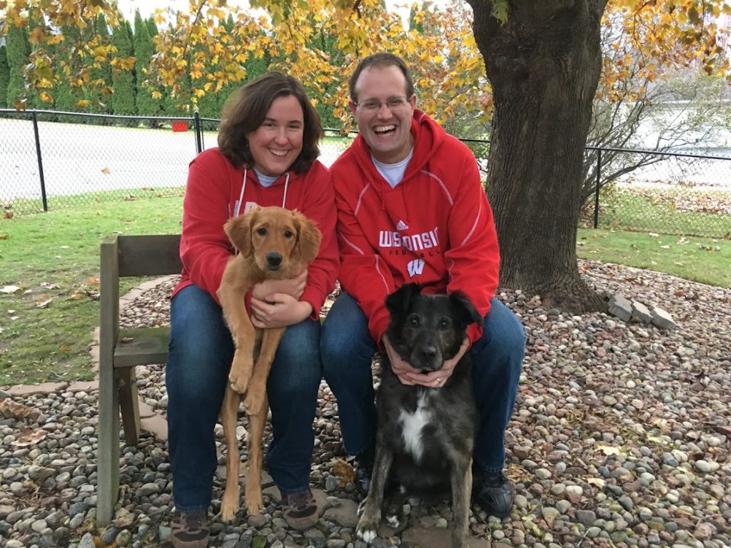 smiling man and woman sitting on bench in red sweatshirts holding two dogs