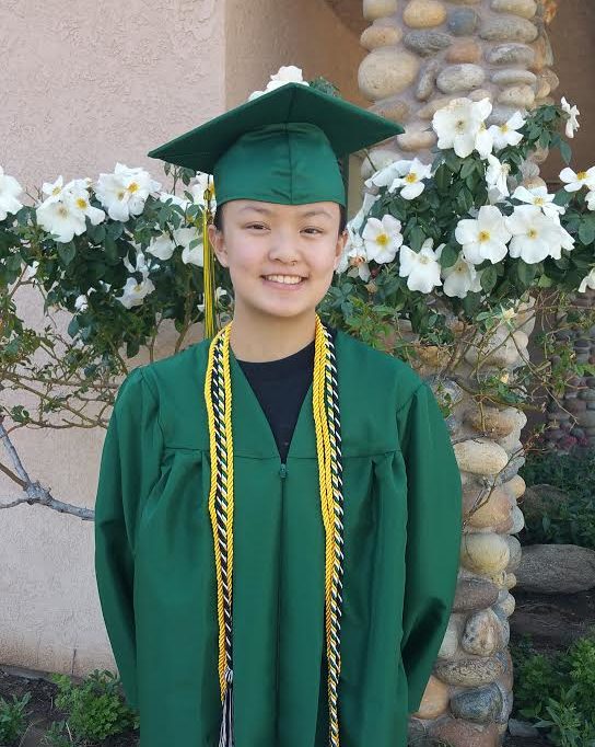 smiling girl standing in front of flowered bush wearing green graduation cap and gown