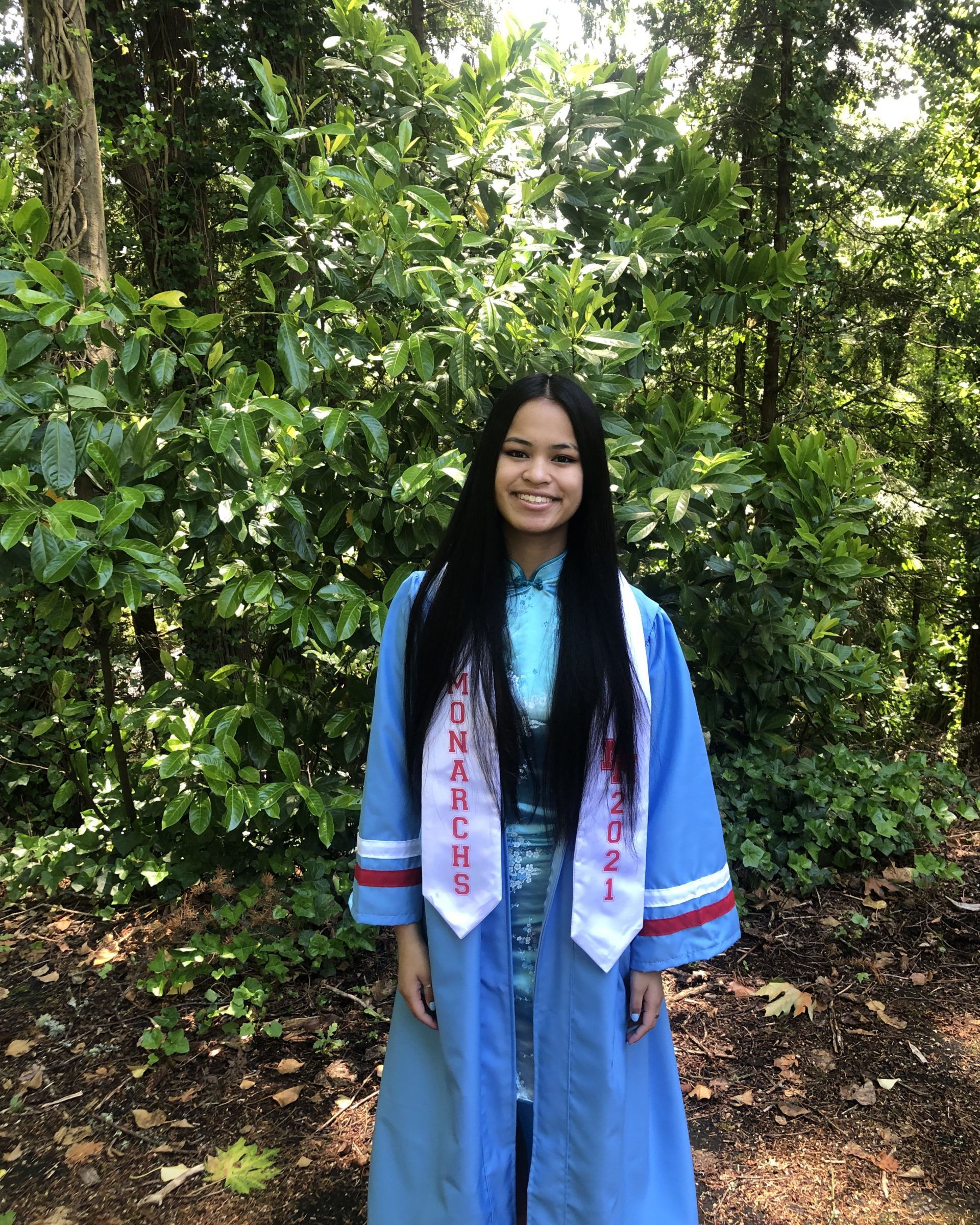 smiling girl in forest wearing blue graduation gown and sash that says "monarchs 2021"