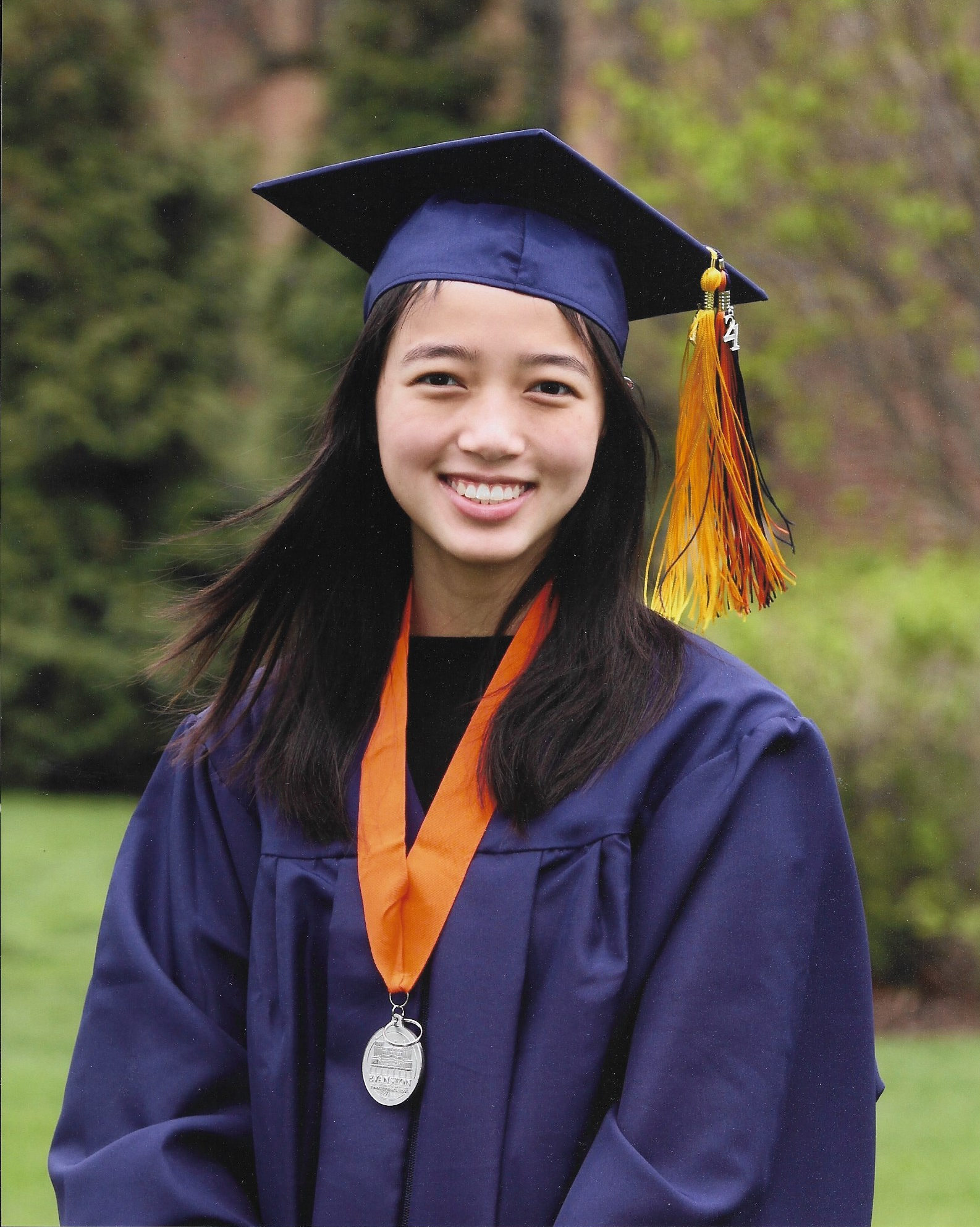 smiling girl standing in field wearing navy blue graduation cap and gown and medal with orange ribbon