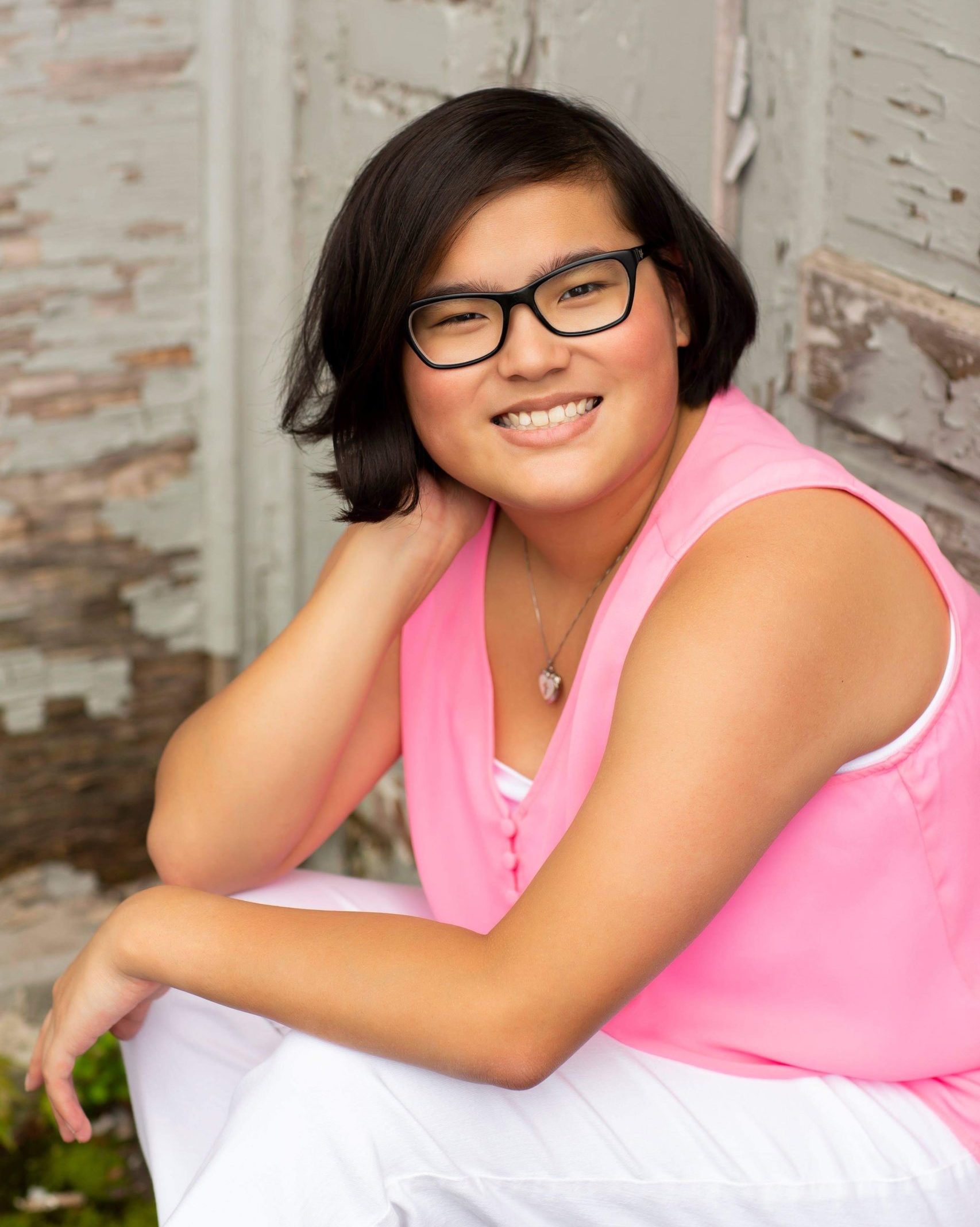 smiling girl sitting with elbows on knees wearing glasses pink tank top and white pants