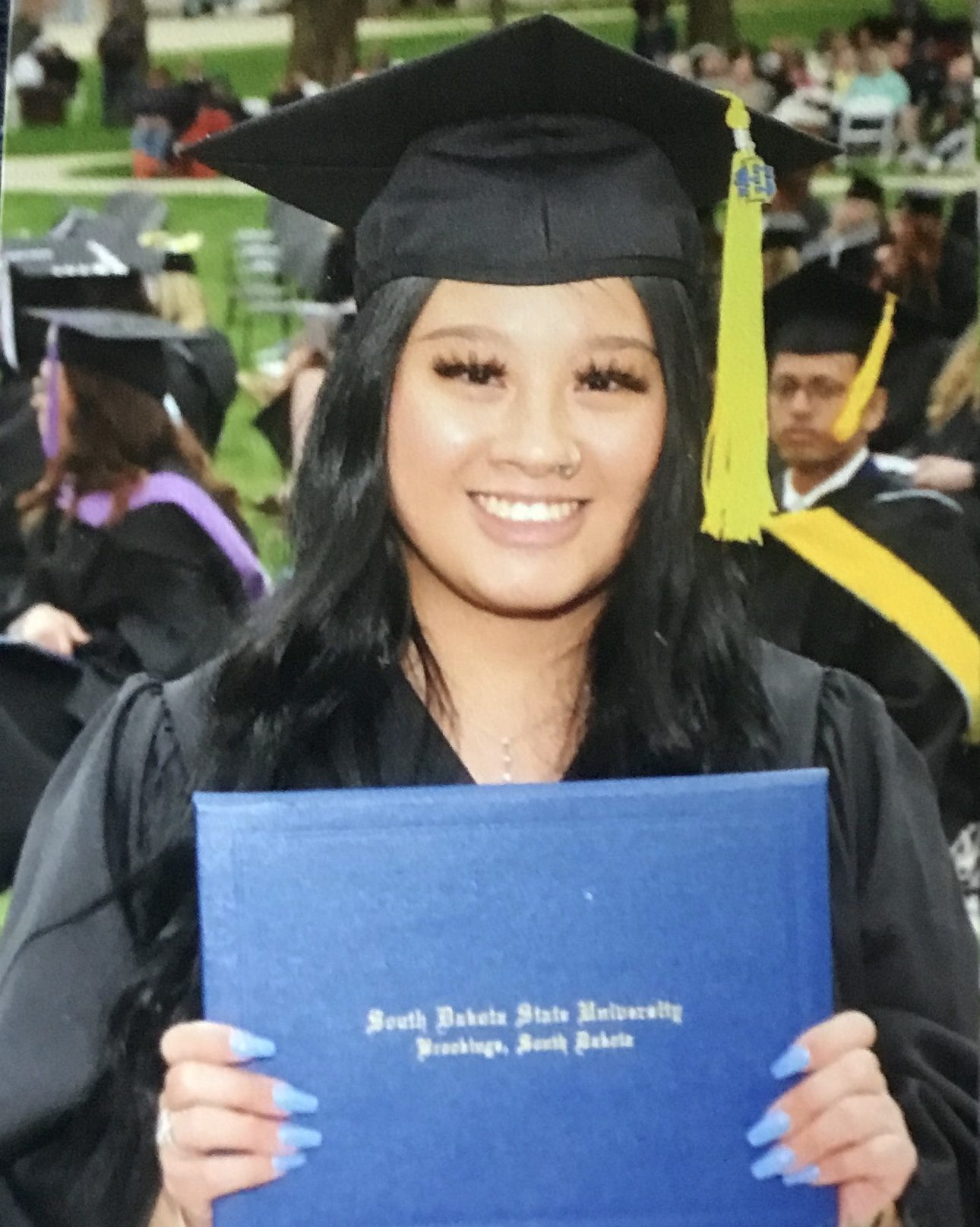 smiling girl wearing black graduation cap and gown and holding blue diploma