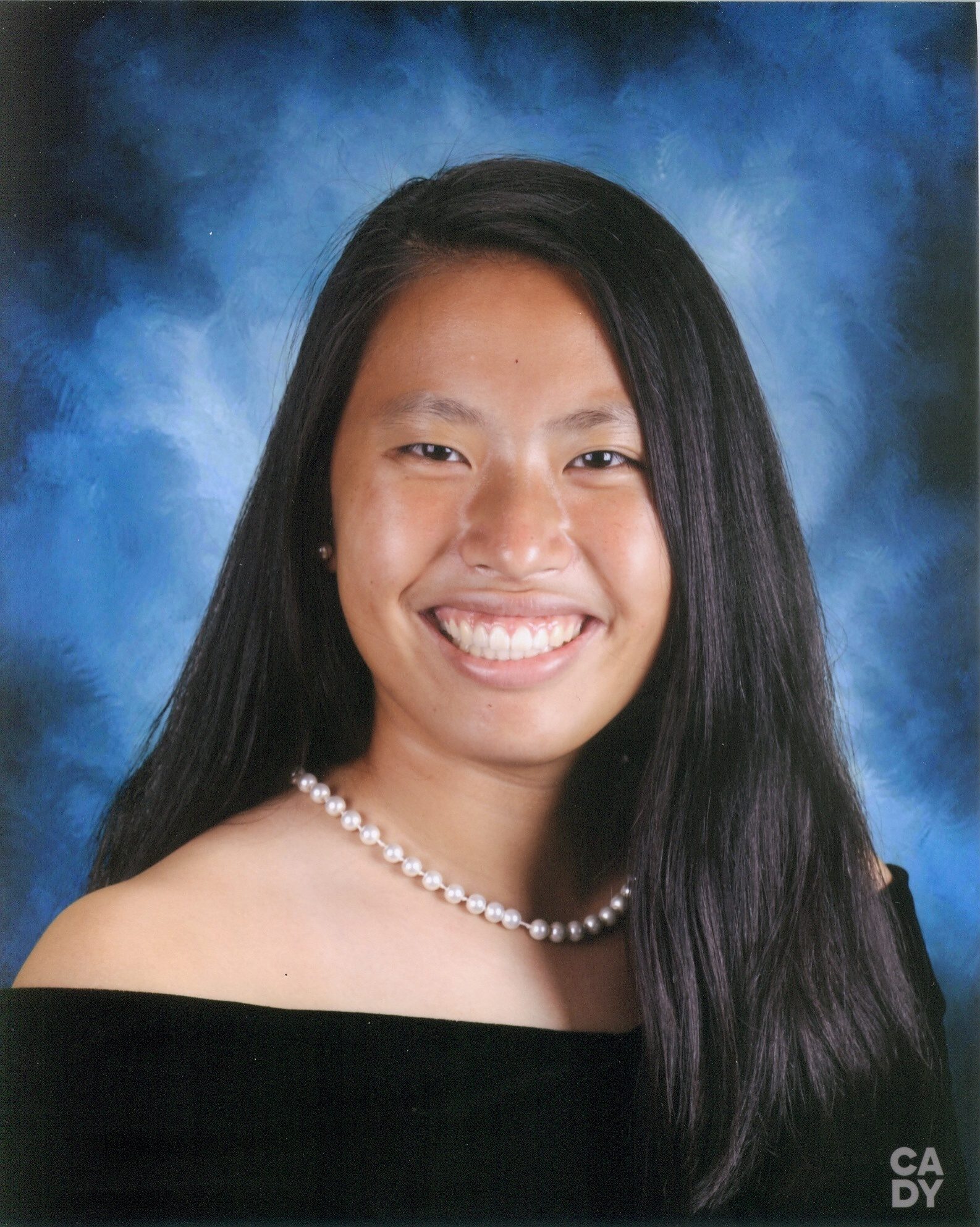 school photo of smiling girl wearing black off the shoulder top and pearl necklace against blue background