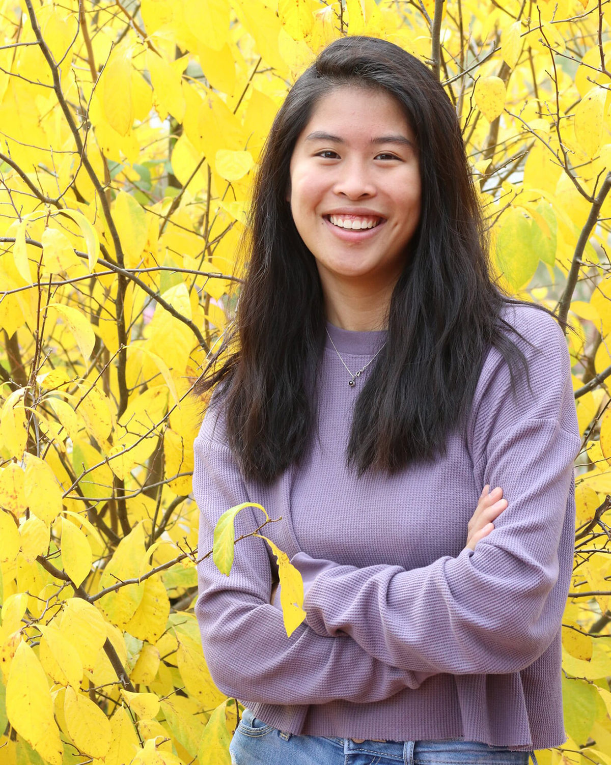 smiling girl wearing lavender shirt standing in front of yellow tree with arms crossed