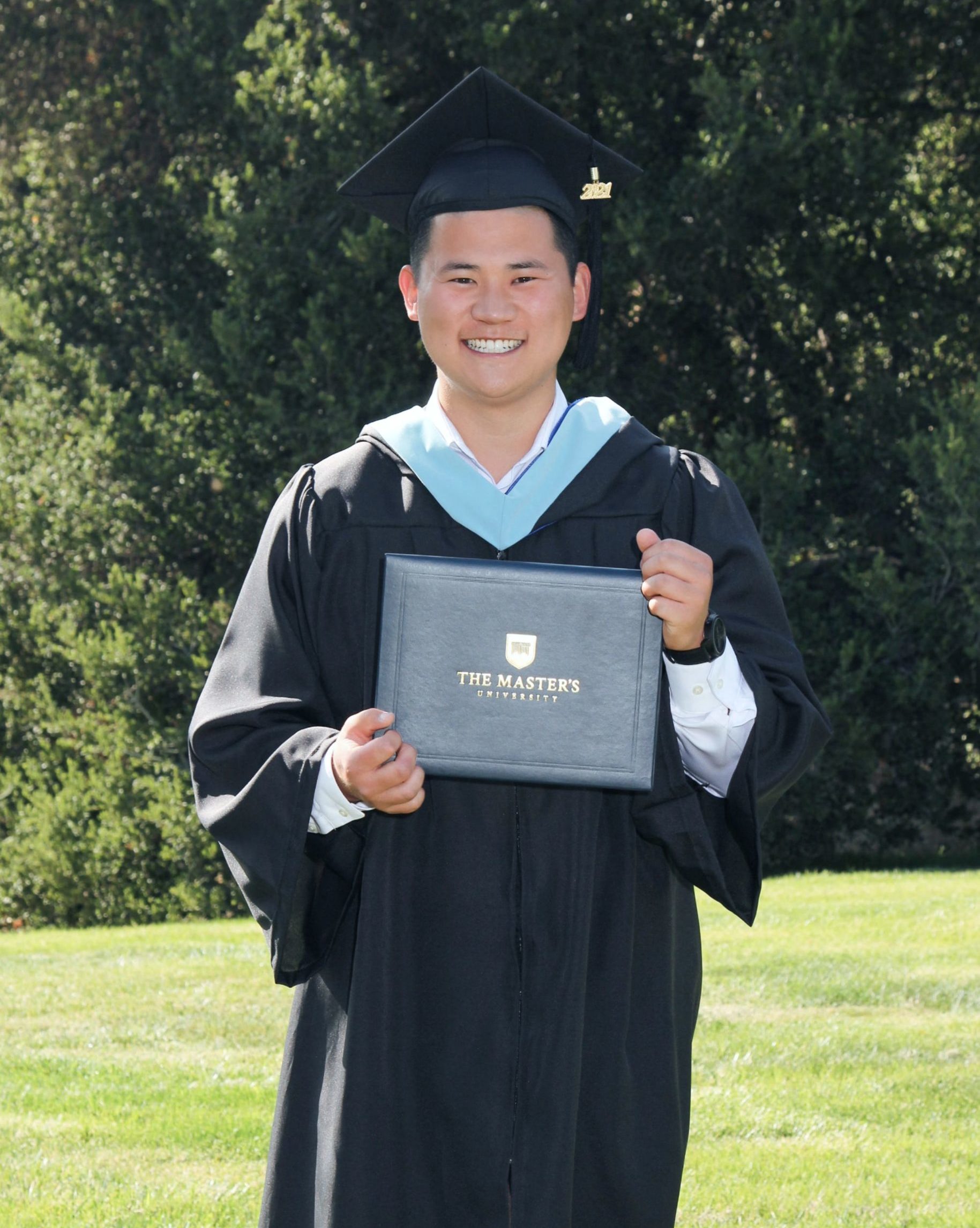smiling man wearing black graduation cap and gown light blue masters cape and holding diploma