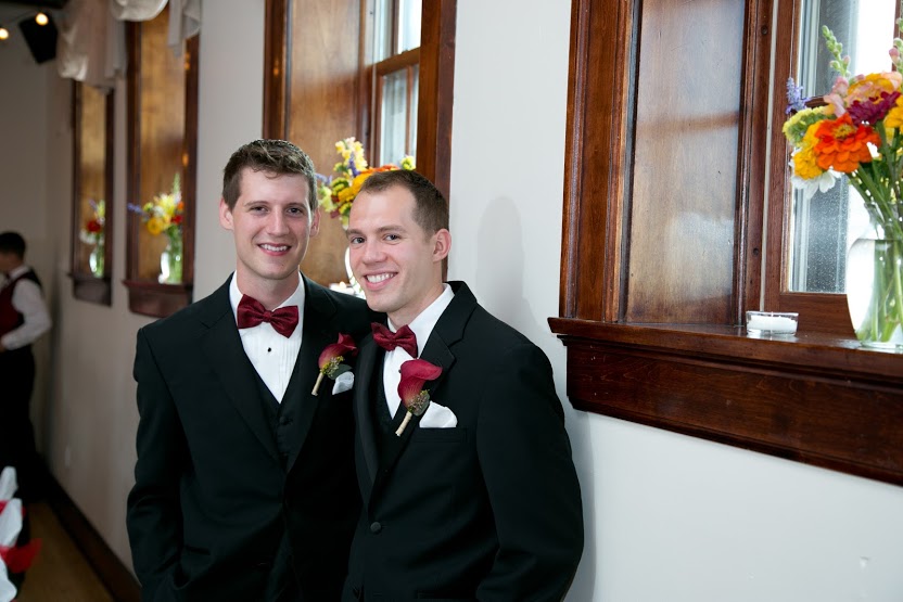 two smiling men in black suits with red bow ties