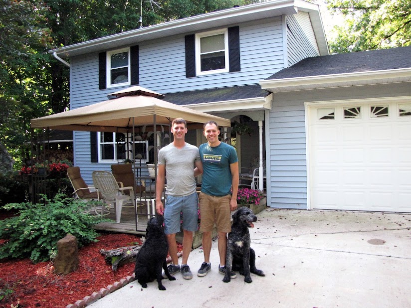two men with arms around each other and two black dogs in front of light blue two story house