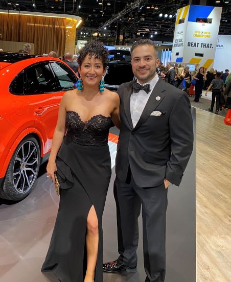 smiling woman in long black dress with smiling man in tuxedo in front of red car
