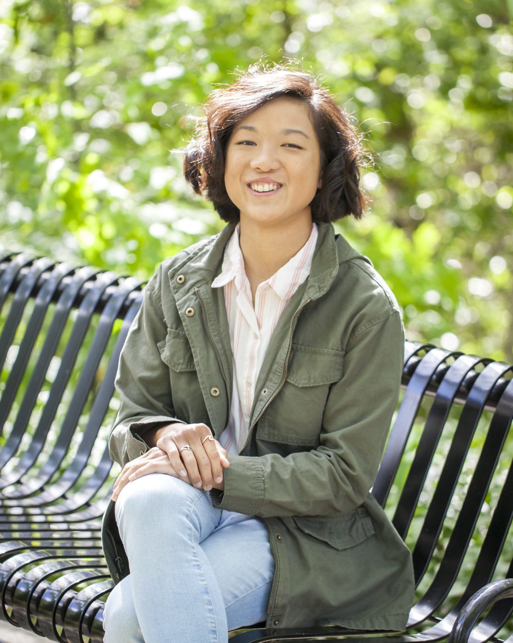 smiling girl with short hair sitting on black metal park bench wearing jeans and green jacket