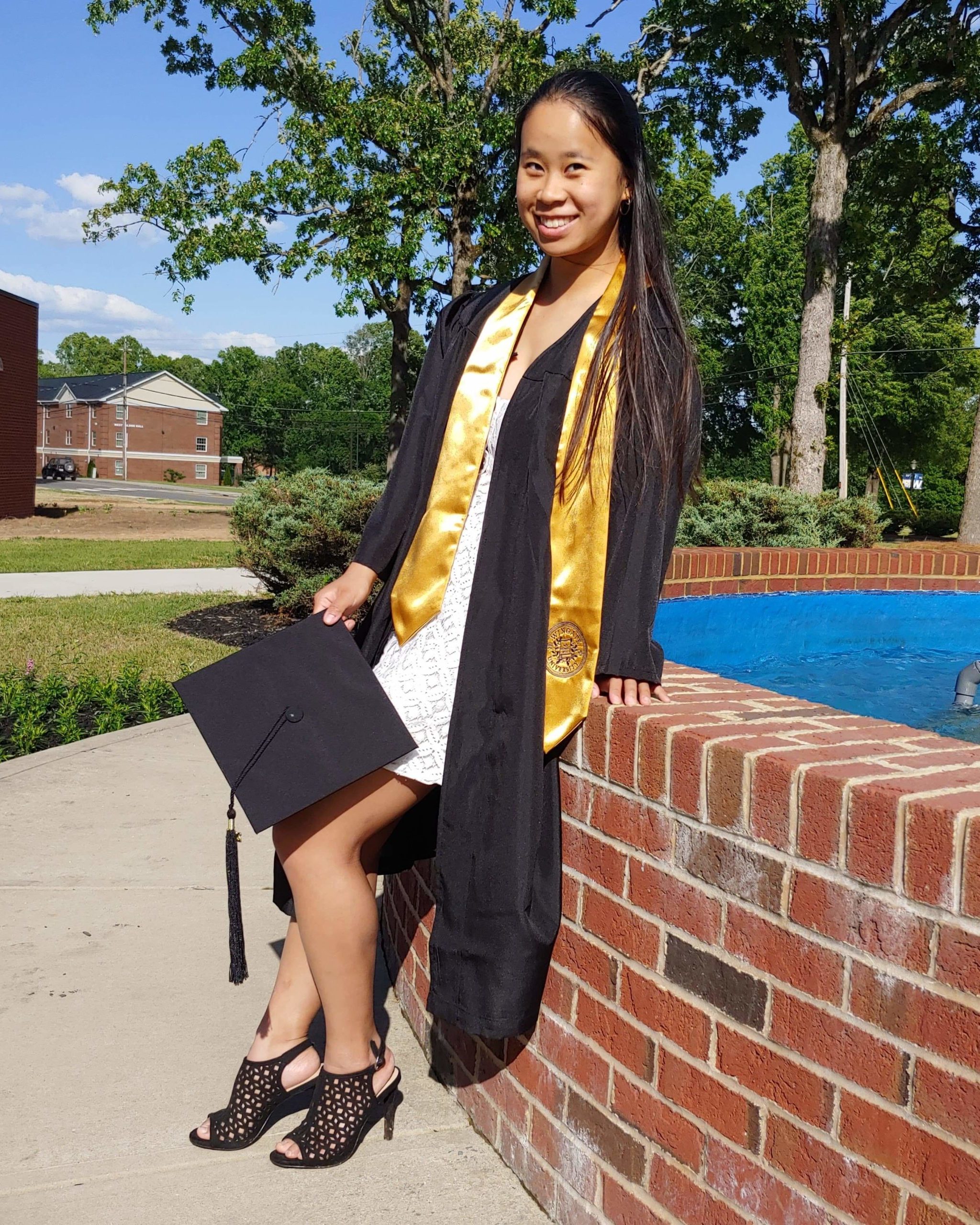 smiling girl sitting on the edge of a brick fountain wearing black graduation cap and gown and yellow sash
