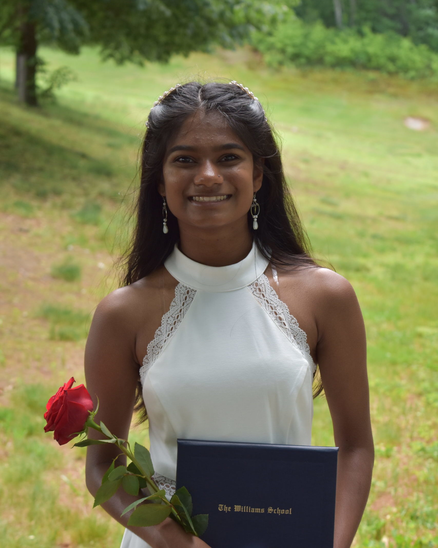 smiling girl wearing white dress and standing in park holding red rose and diploma