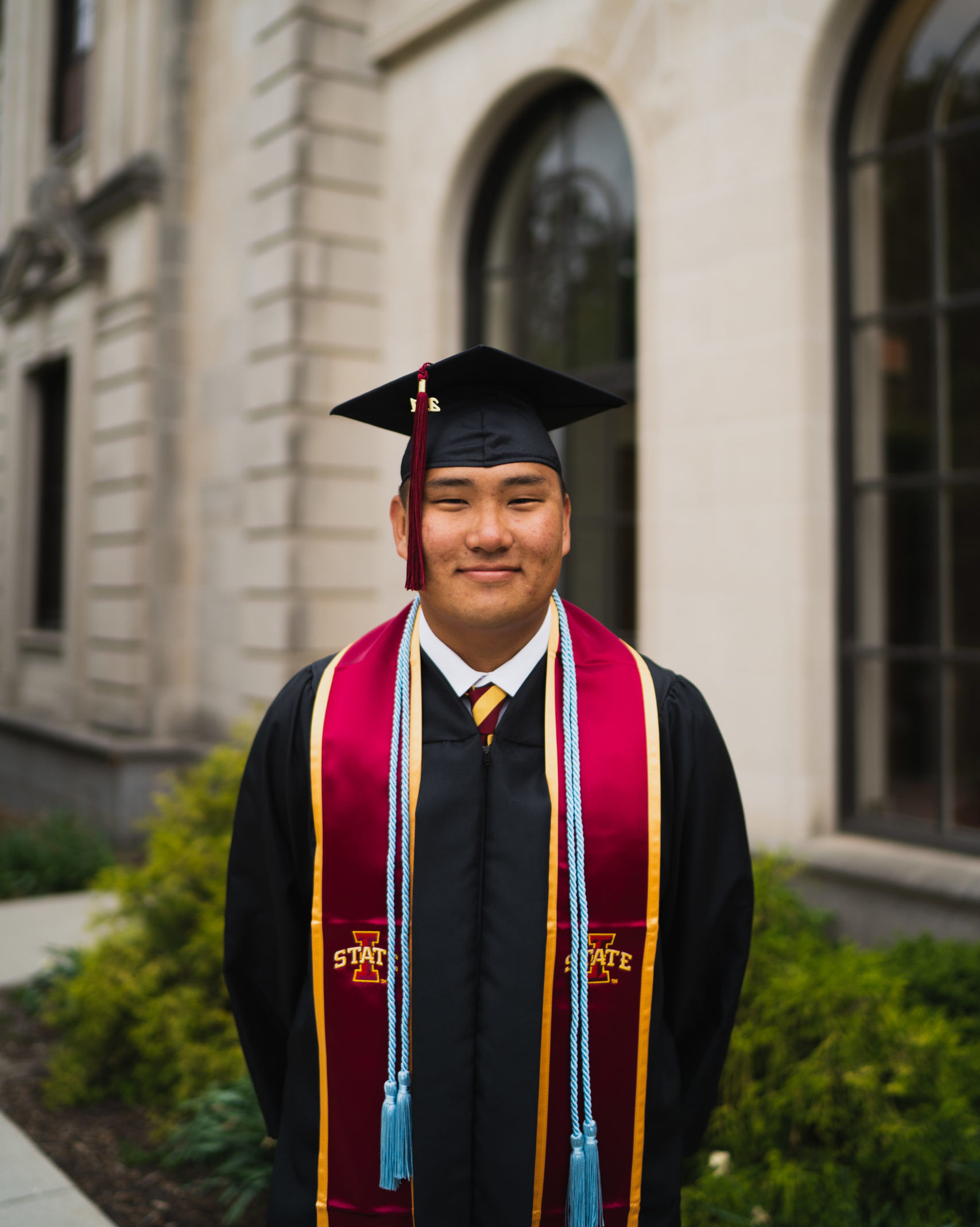 smiling man wearing black graduation cap and gown and burgundy sash