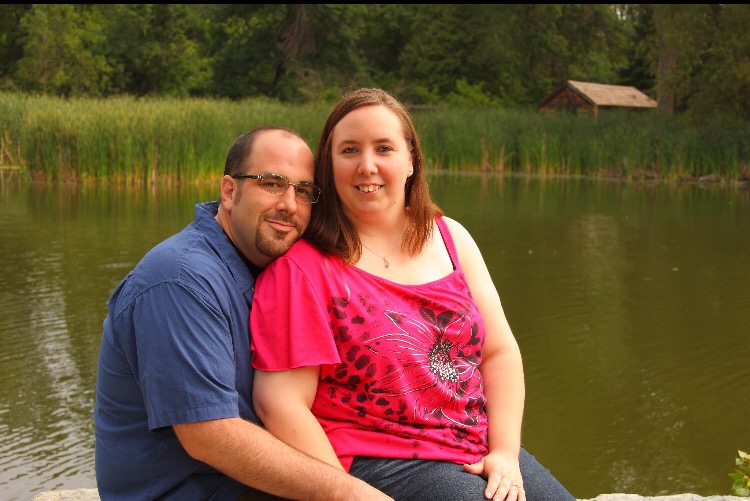 smiling woman in pink tank top sitting on smiling man in blue shirt in front of a lake
