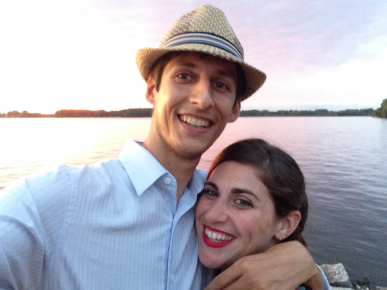 man in fedora with his arm around woman in red lipstick in front of a lake