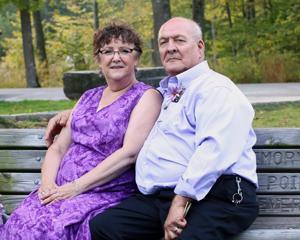 smiling woman in purple dress next to man in purple shirt on a park bench