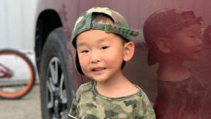 smiling little boy adopted from Korea wearing camo shirt and hat in front of red car