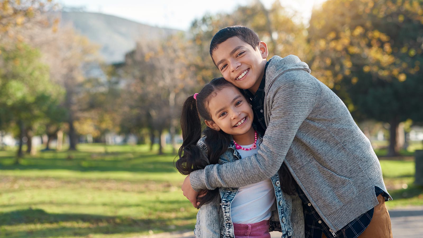 smiling older brother with arms around smiling younger sister in a park