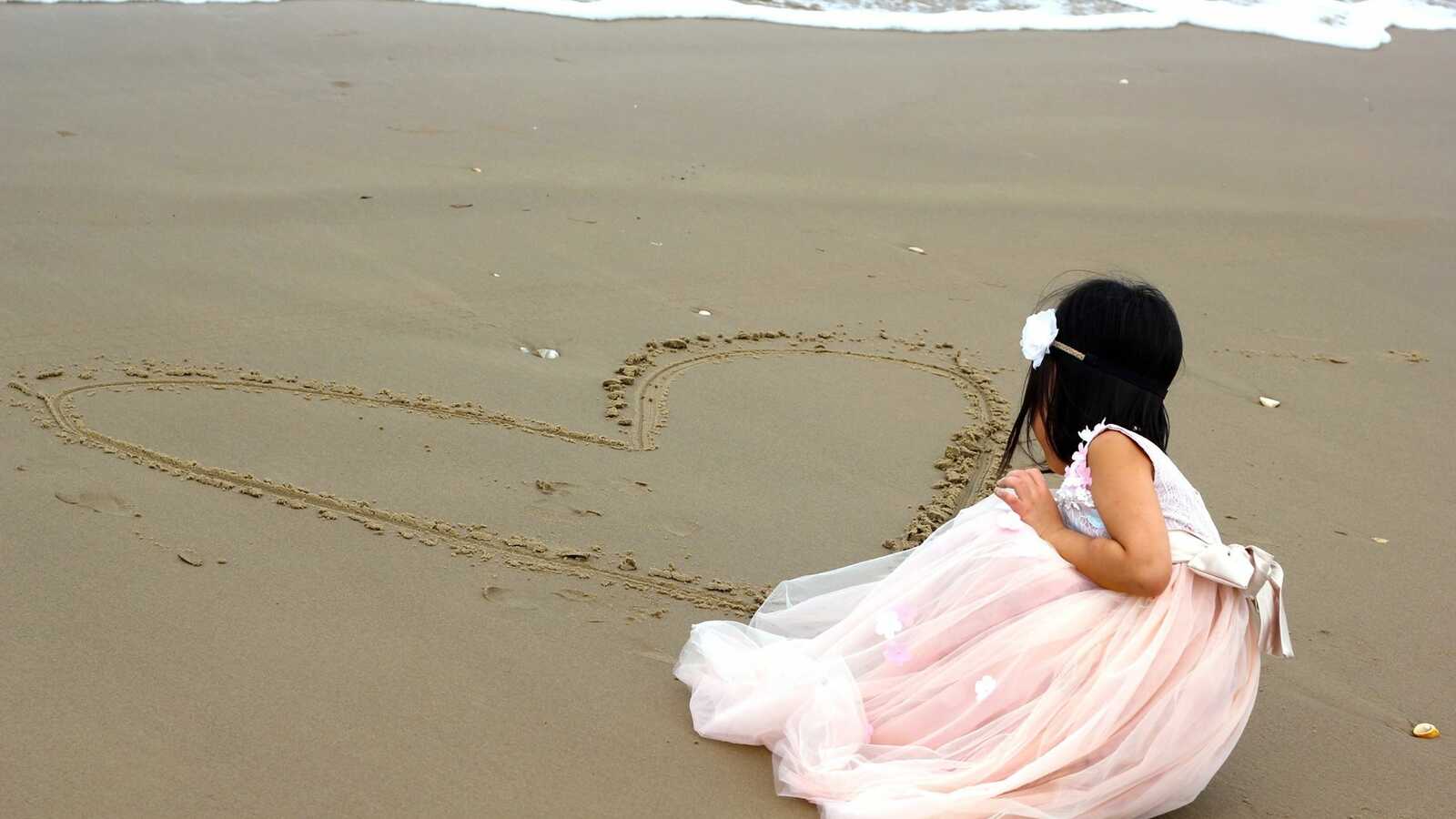 girl draws heart in sand by ocean