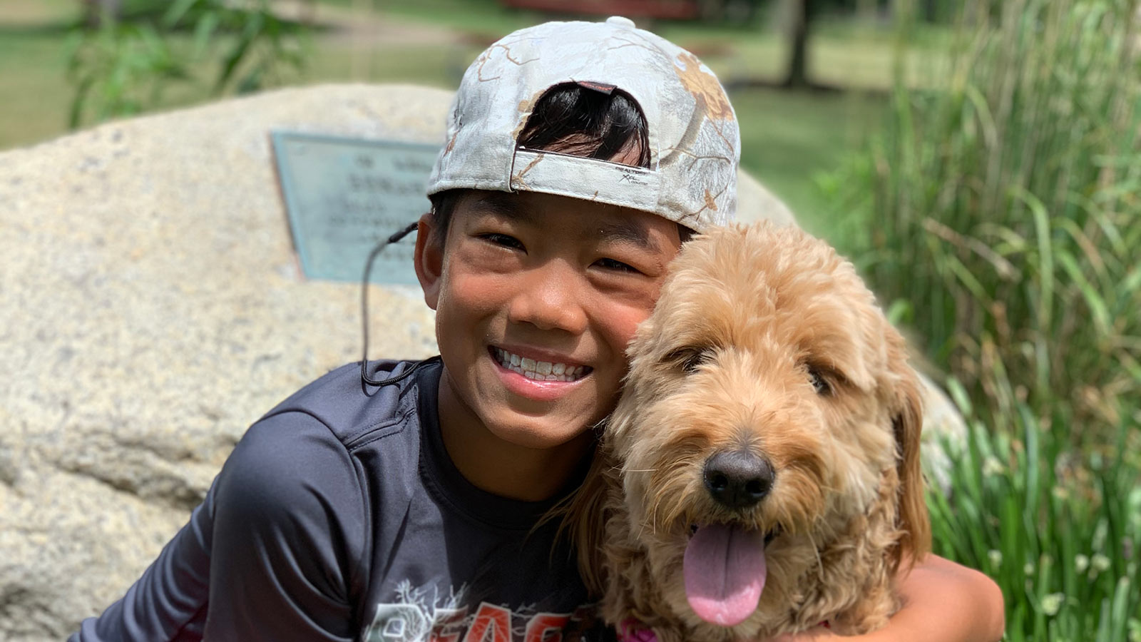 smiling boy adopted from vietnam wearing backwards hat with arms around doodle dog