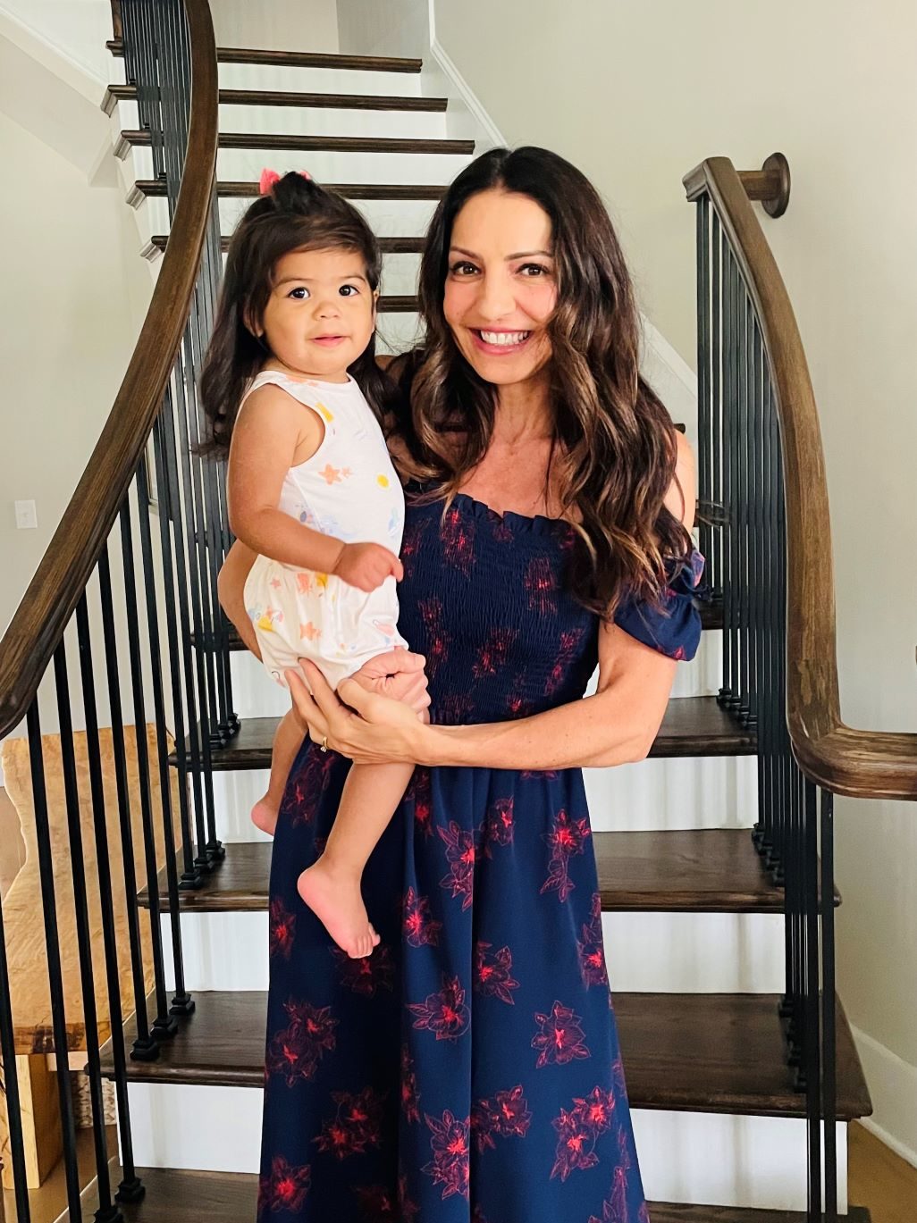 smiling woman in navy blue dress holding toddler girl in front of staircase