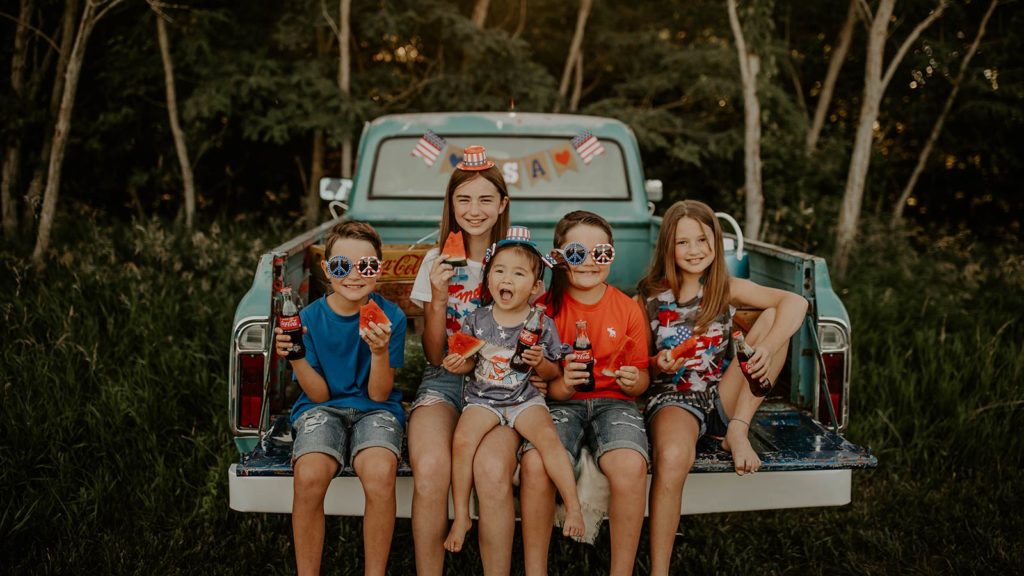group of siblings and international adoptee from china sitting in bed of blue truck wearing fourth of july shirts and glasses