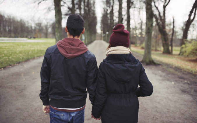 Rear view of teenage couple walking in park holding hands wearing warm fall clothes.