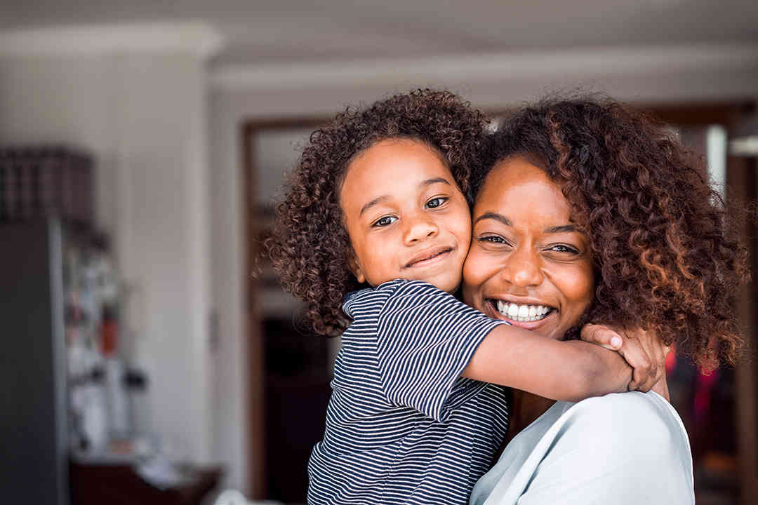 Mother and young child standing in kitchen smiling and hugging with child's arms wrapped around mother's neck tightly with their faces touching