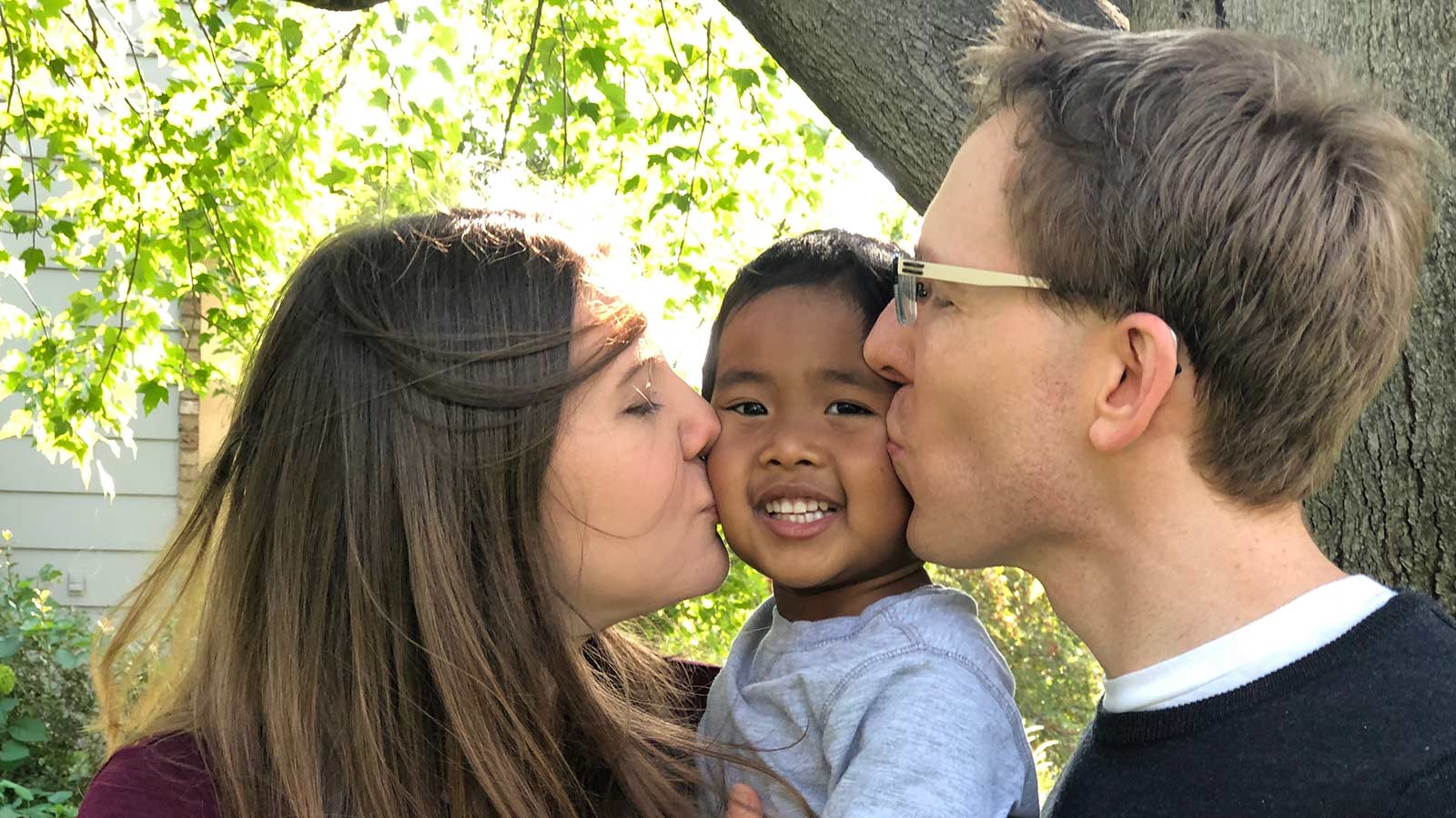 adoptive parents giving son adopted from Thailand a kiss on the cheek