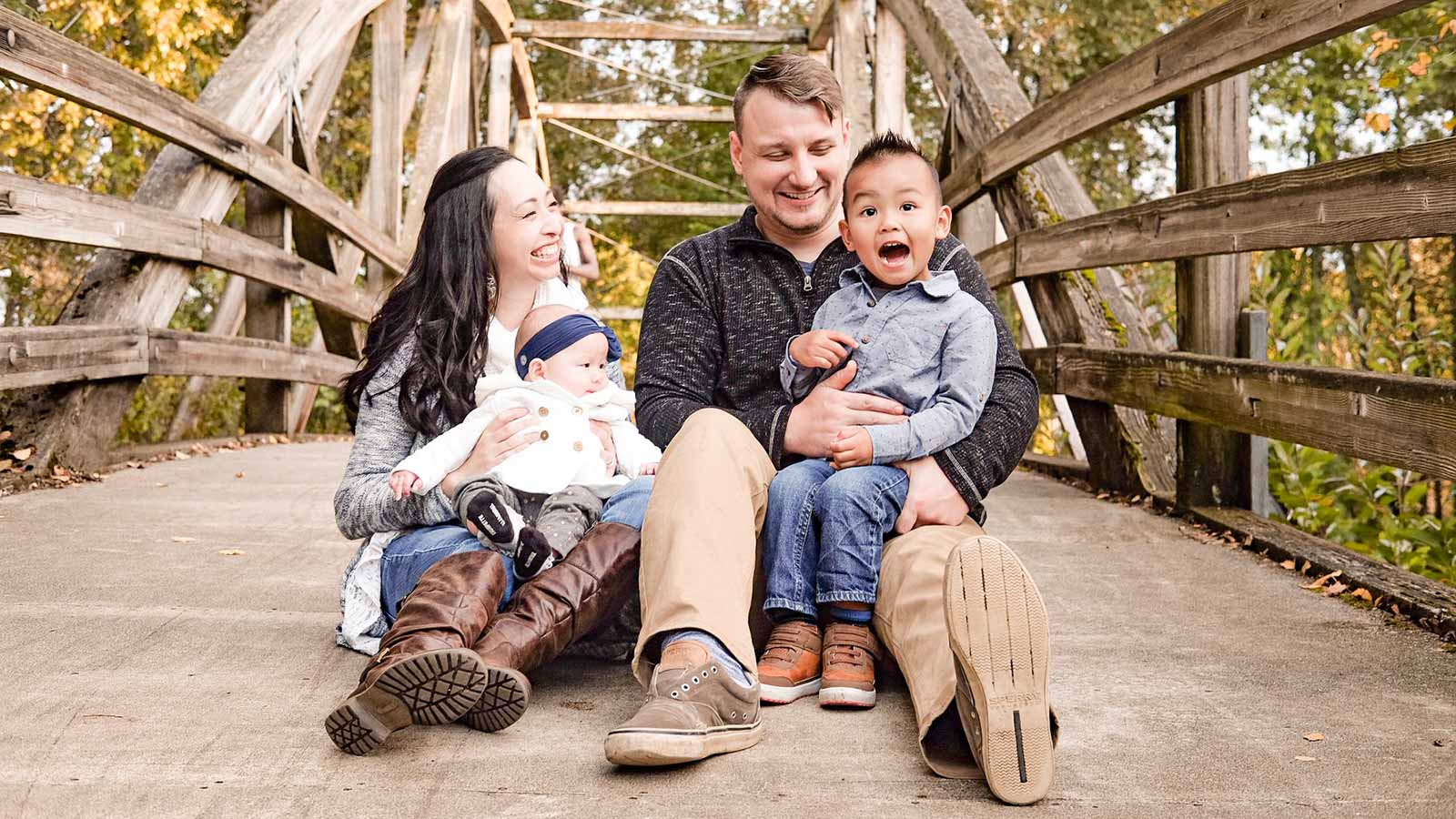 adoptive parents with son adopted from thailand and baby sitting on bridge