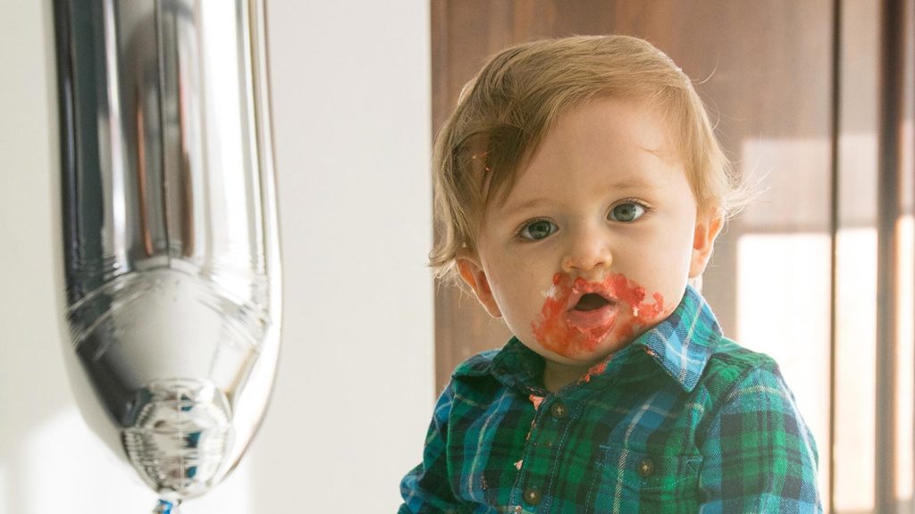 baby with frosting on his face sitting next to balloon