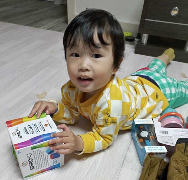 Little boy laying on tummy on the ground playing with toys looking up at camera happily.