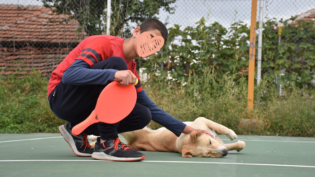 A young boy holds a tennis racket and pets a dog